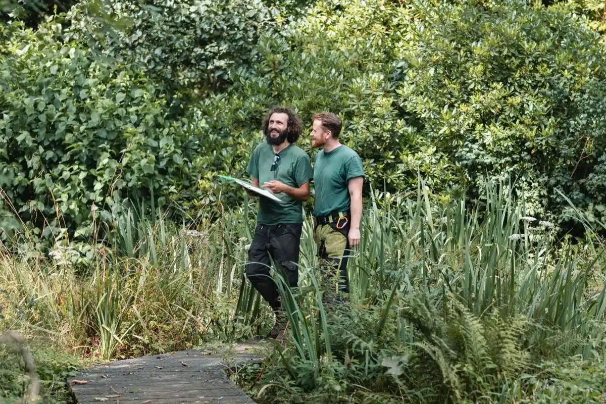 Inside a greenhouse, a man and woman pose among lush greenery and vibrant plants.