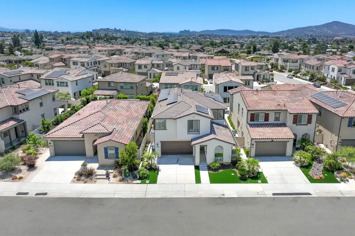 Aerial view of a suburban neighborhood with rows of similar two-story homes featuring red-tiled roofs. Solar panels are visible on some rooftops, and manicured lawns line the street. Background shows distant hills under a clear blue sky.