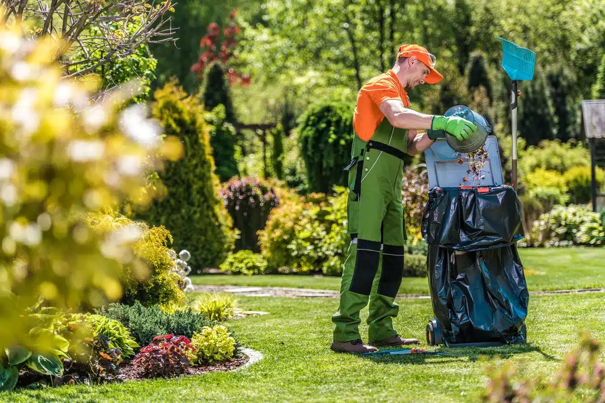 A gardener in orange and green work attire uses a leaf shredder in a lush, vibrant garden under sunlight. The scene conveys a peaceful, industrious mood.
