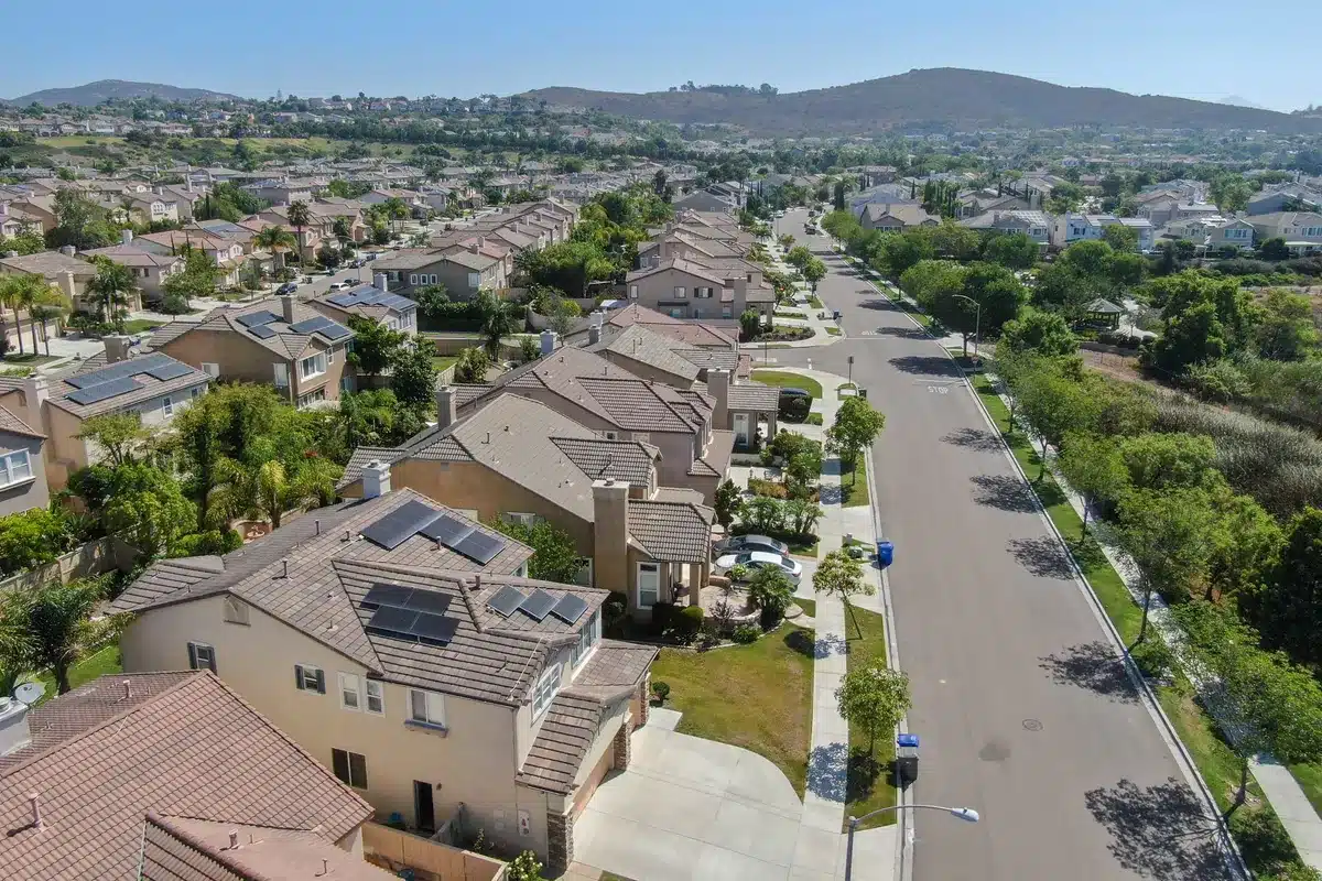 Aerial view of a suburban neighborhood with rows of beige and brown houses featuring solar panels. Tree-lined streets and hills in the background under a clear blue sky.