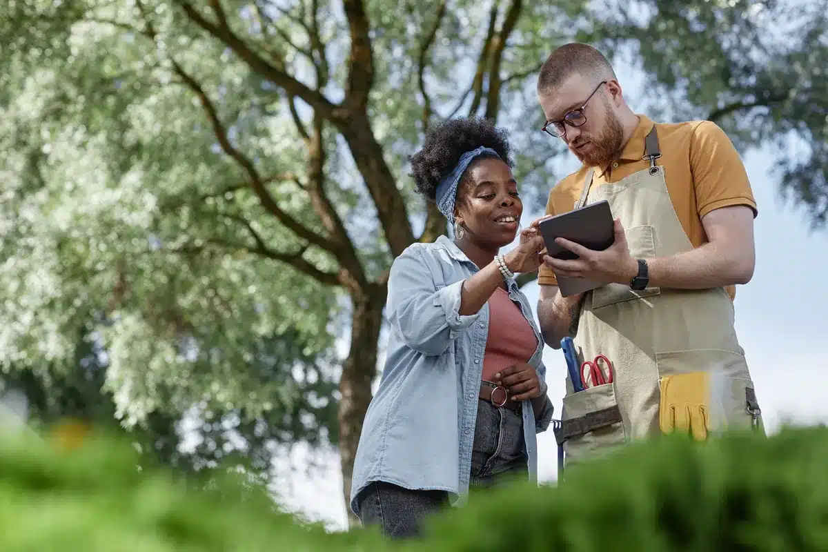 A woman and man in a garden study a tablet, surrounded by green foliage. The woman smiles while pointing at the screen, conveying a collaborative mood.