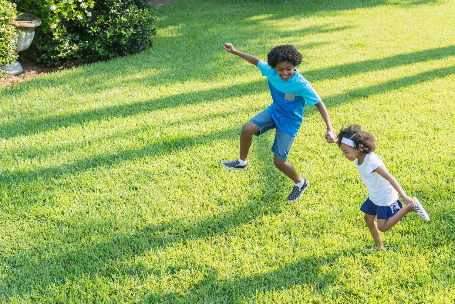 Two children joyfully run and jump on a sunlit lawn. The older child wears a blue shirt and shorts, the younger a white shirt and navy skirt. Lush greenery surrounds them.