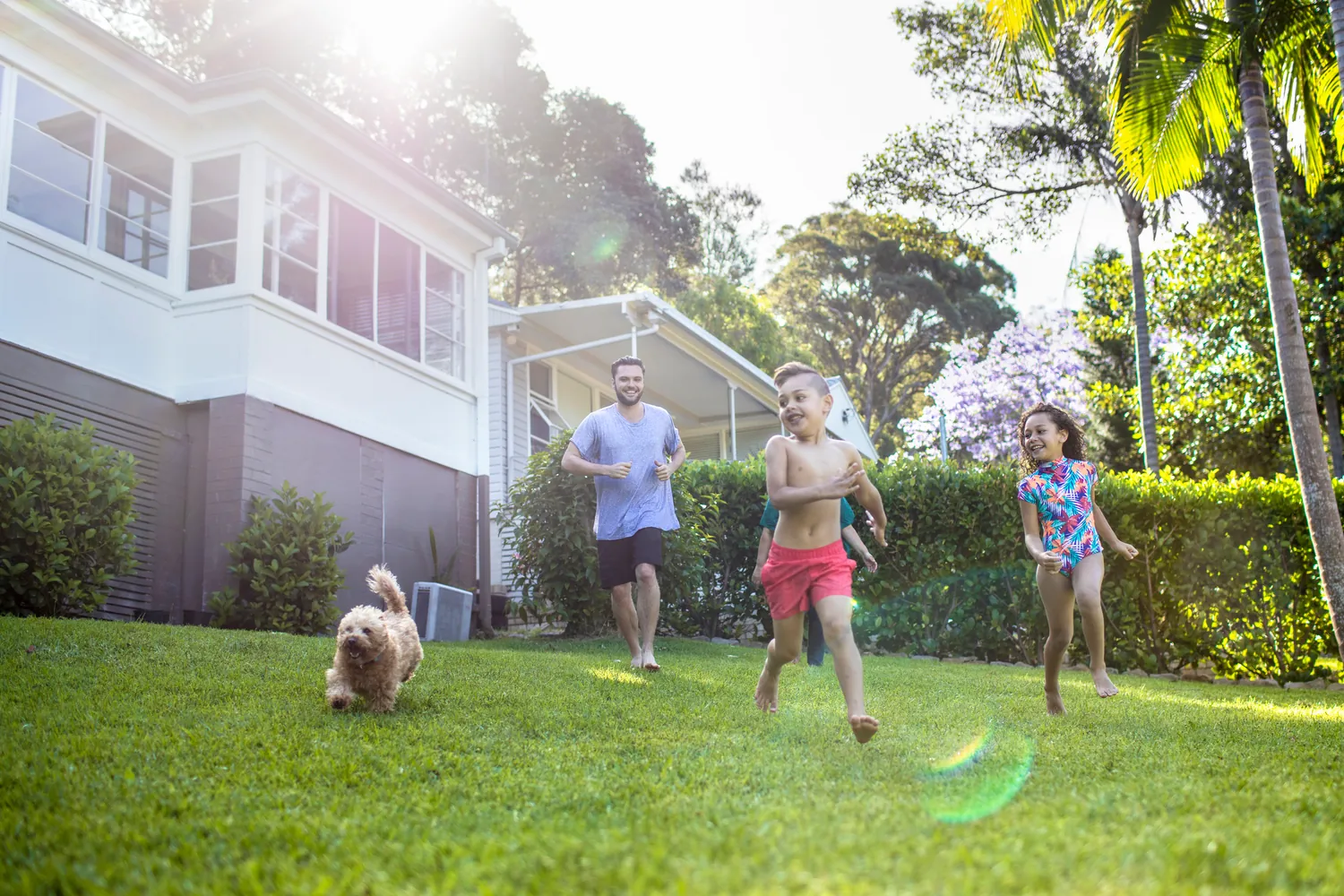 A joyful family scene with a man, two children in swimsuits, and a dog running on a sunny lawn in front of a house, exuding energy and happiness.