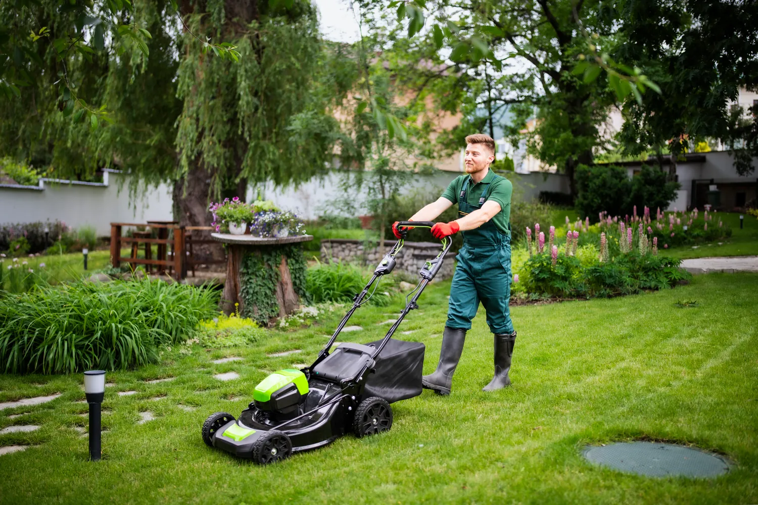 A person mows a lush green lawn with an electric mower, surrounded by vibrant garden plants and trees. The scene feels peaceful and tidy.
