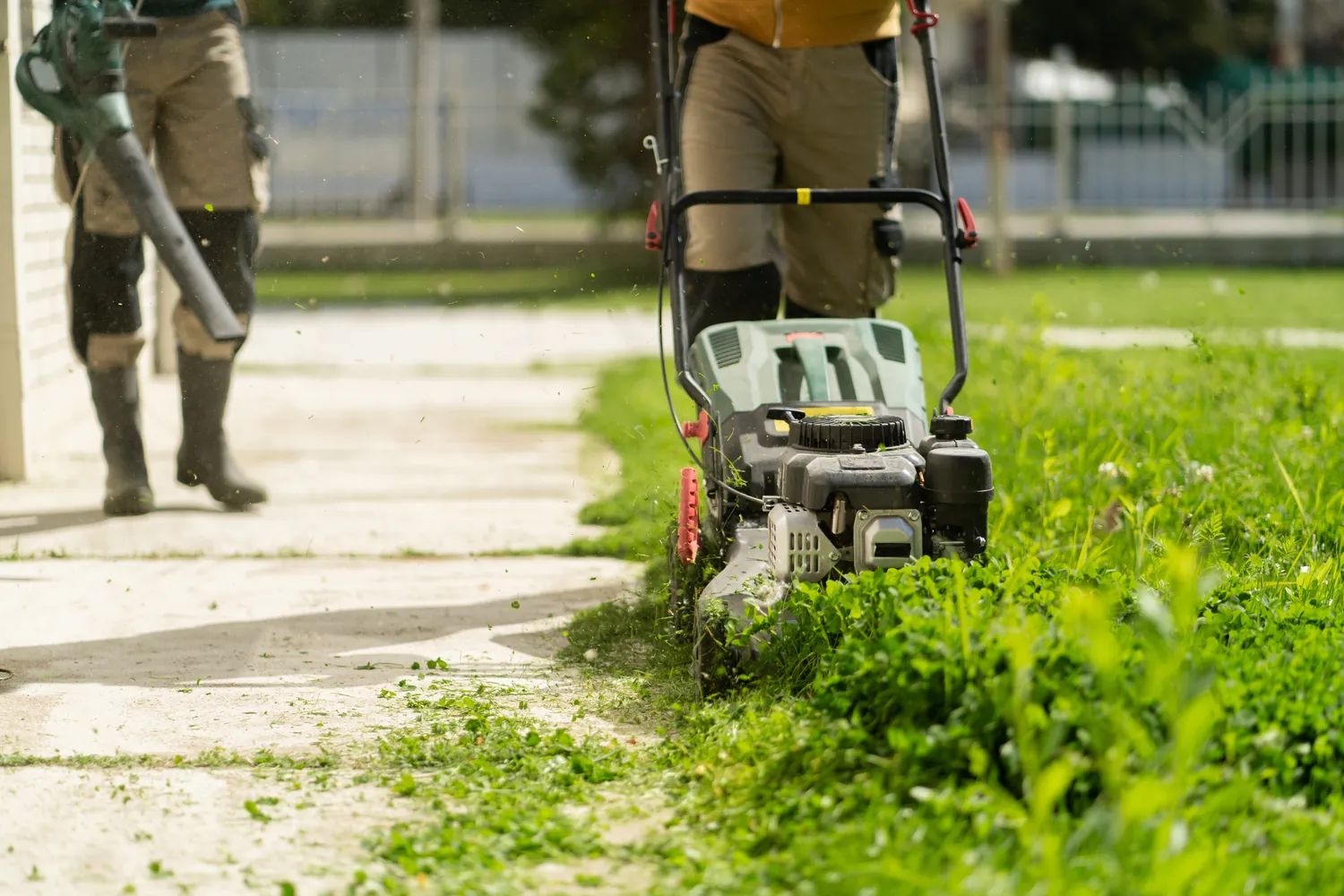 Two people are tending a lawn; one operates a lawnmower cutting grass, while the other uses a leaf blower. The setting conveys a sense of outdoor maintenance.