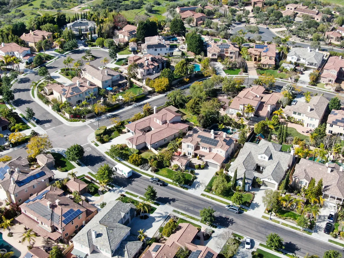 Aerial view of a suburban neighborhood in bright daylight featuring a FedEx delivery truck driving on a residential street lined with houses and trees.